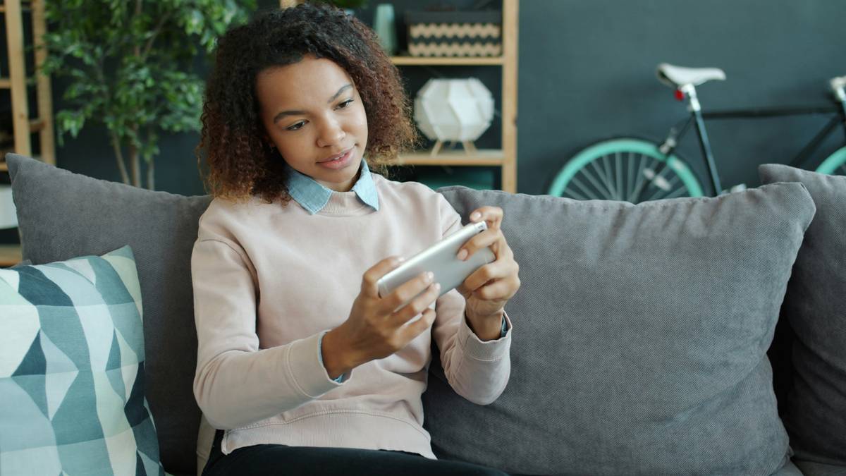 Happy African American girl is playing smart phone game enjoying entertaining activity touching screen smiling at home. People and hobby concept.