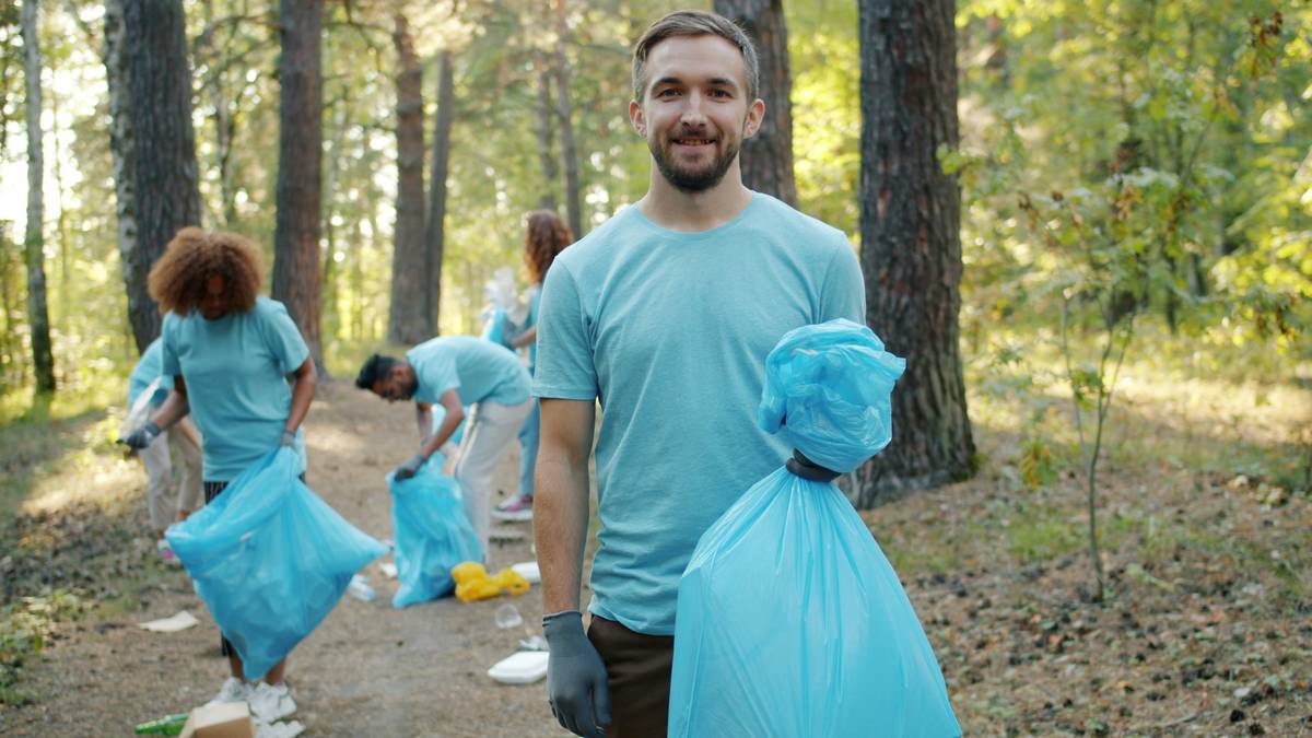 Member of volunteering team attractive guy in uniform holding bin bag with trash in forest smiling looking at camera while group of youth collecting garbage