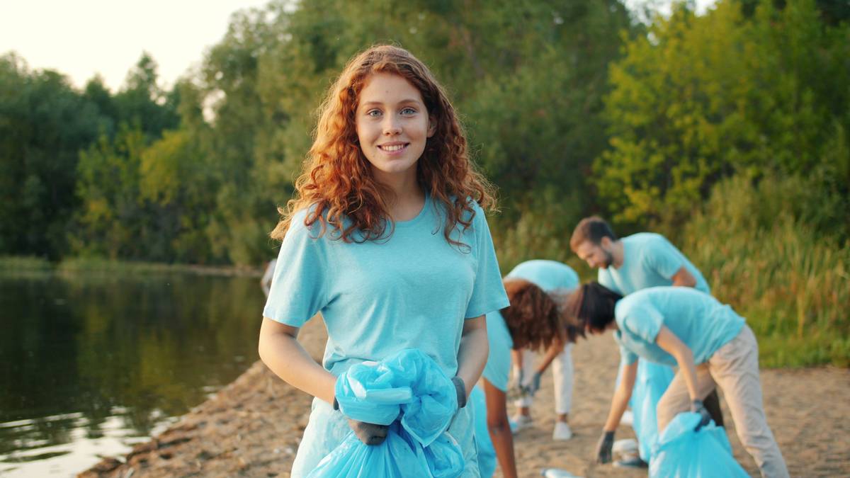 Portrait of beautiful girl female volunteer on beach looking at camera smiling while multi-ethnic team is cleaning lake shore gathering garbage in bin bags.