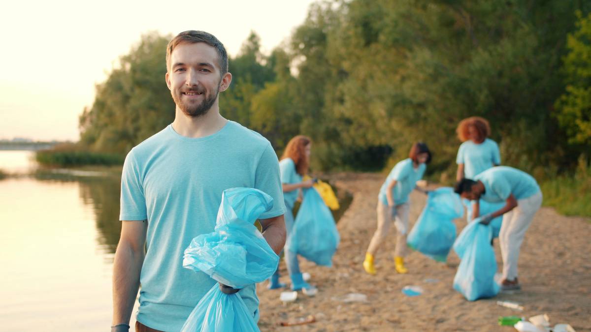 Slow motion portrait of young man volunteer standing on contaminated river bank holding trash bag looking at camera smiling while people are working in background.